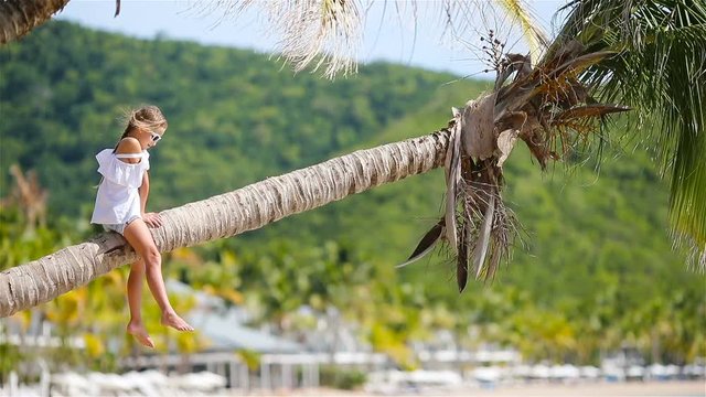 Little girl at tropical beach sitting on palm tree and havinf a lot of fun. Kid on caribbean vacation in Antigua island