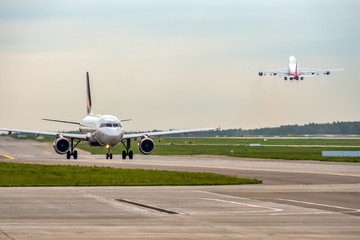 Modern passenger airplane taxiing while big widebody aircraft taking off at airport.