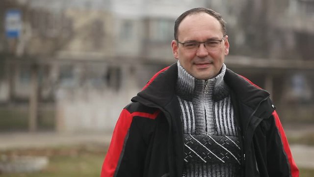    A cheerful view of a middle-aged man in glasses and a sportive zipup jaket smiles optimistically on a city street on a sunny day in winter.