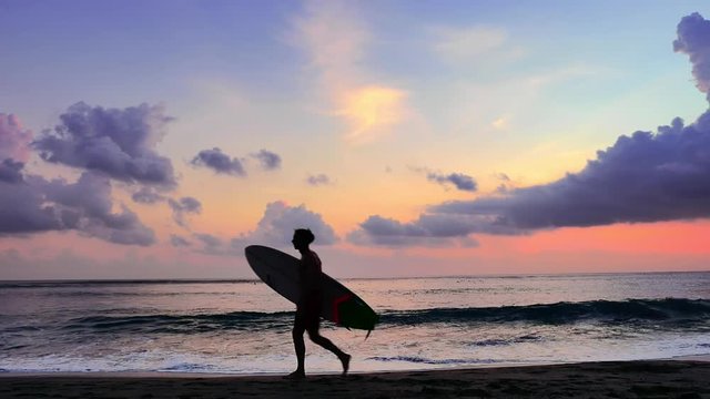 Beautiful slow motion view of male surfer silhouette walking along tropical sea beach at sunset holding surf board. Vacation background of Bali island, Indonesia