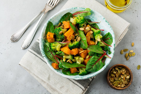 Salad with a baked pumpkin, chard, broccoli, and pumpkin seeds in ceramic plate on stone or concrete background table background. Selective focus. Rustic style. Top view. Copy space.