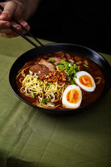 Japanese ramen with pork belly, mushrooms and marinated eggs on green linen tablecloth. Male hand holding chopsticks