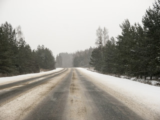 Straight dirty asphalt road with snow roadside surrounded with trees