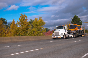 Fototapeta na wymiar Big rig semi truck transporting equipment on step down semi trailer moving on wide autumn highway