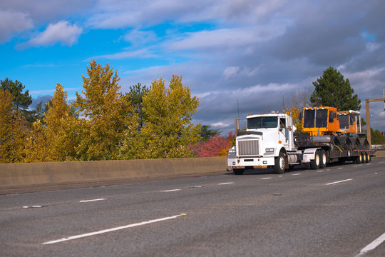 Big rig semi truck transporting equipment on step down semi trailer moving on wide autumn highway
