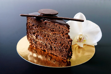 A piece of chocolate cake with chocolate cream on a golden napkin with a white orchid flower on a black background.