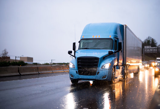 Modern big rig blue semi truck with trailer driving on rainy wet highway with headlight reflection