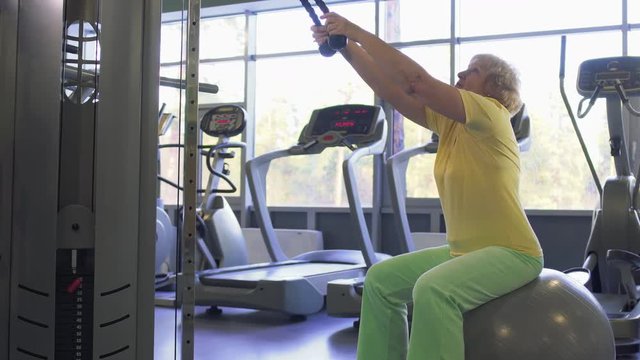Elderly woman makes pull up exercise on training apparatus in the gym