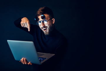 Bearded man in flip-up glasses looking at camera with excitement holding laptop on black background.