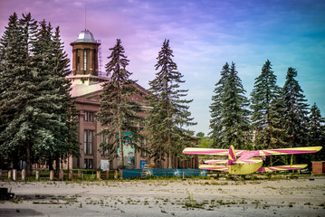 Obraz premium An old yellow and pink plane in an abandoned airport next to dark forest in an overcast day. Military plane, old russian aircraft. Soviet mass-produced single-engine biplane at an field aerodrome