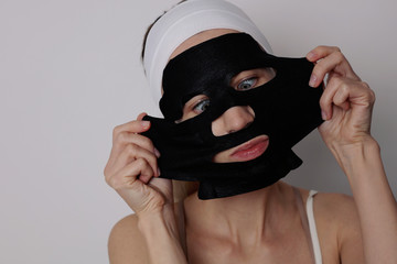 Young woman applying black charcoal cosmetic facial mask and making funny face. Skin care, beauty treatments.