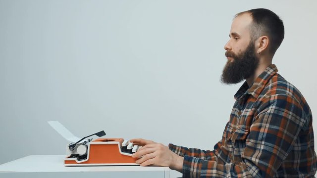 Side view of hipster man with red vintage typewriter sitting at table looking forward at blank copy space, looking at camera smiling gesturing thumb up