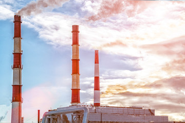 Tubes of factory on a background of the cloudy sky. Through the clouds the sun is visible.