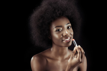 Obraz premium smiling african american woman applying lipstick and looking at camera isolated on black