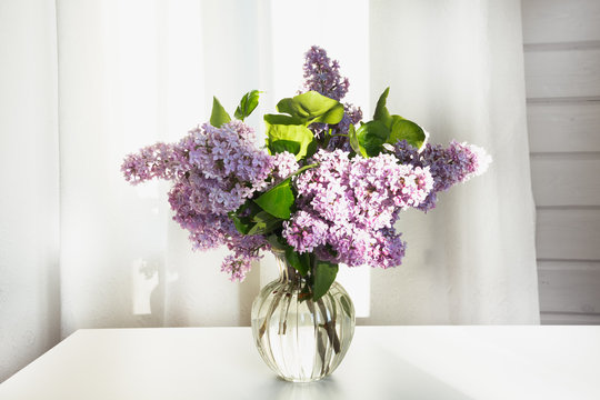 A bouquet of fresh lilacs in a beautiful glass vase on a white table in a farmhouse.