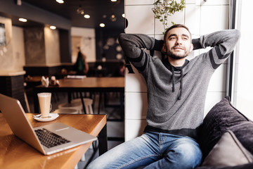 Fototapeta premium Relaxed young man holding hands behind head and smiling while sitting at the table with laptop in modern cafe