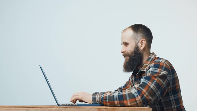 Bearded hipster man sitting at table working with laptop, turning to camera and gesturing thumb up, side view