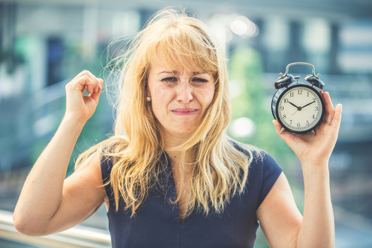 busy caucasian businesswoman messup hair and stress emotion hand hold black alarmclock with blur bacgrund of urban downtown city