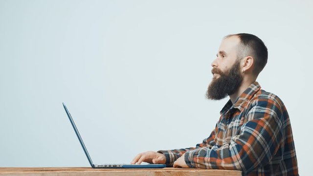 Bearded hipster man sitting at table with laptop, looking at blank copy space in front of him, side view