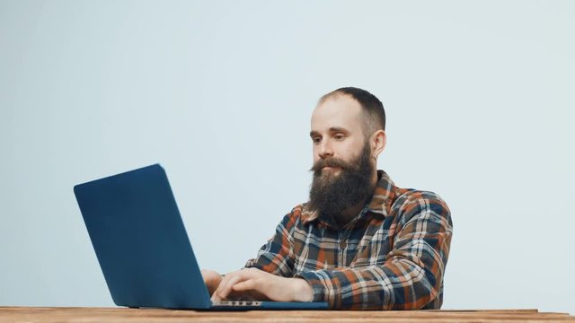 Bearded hipster man sitting at table working with laptop, turning to camera and gesturing thumb up
