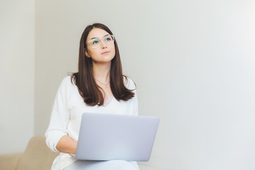 Naklejka premium Pensive young brunette female sits in front of opened laptop, thinks about interesting presentation, wants to impress everyone by her work, wears spectacles, sits against white wall