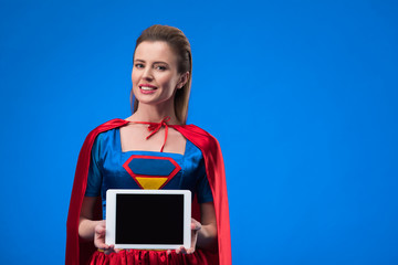 portrait of smiling woman in superhero costume showing tablet with blank screen isolated on blue