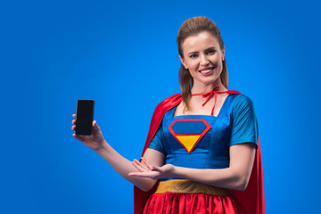 portrait of smiling woman in superhero costume showing smartphone with blank screen isolated on blue