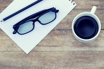 Top view of americano coffee in white cup with white paper, pencil and glasses on wooden background. Workplace and copy space design. Vintage tone.