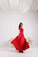 Elegant woman in long red dress standing in white room, dress flashing, flying. Copy space. European tall girl with makeup. Beauty lifestyle concept.