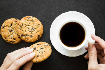Woman's hands holding a cup of black coffee and a cookies with chocolate pieces on the dark table. Resting and enjoying time with coffee and sweets. Drink and snack concept.