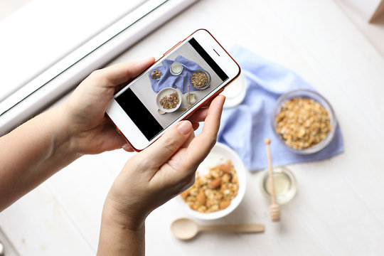 Close up of female hands holding mobile phone taking a picture of granola cereals, milk yogurt and trail mix vegetarian dieting breakfast for a new food blog article. Top view, overhead, copy space.