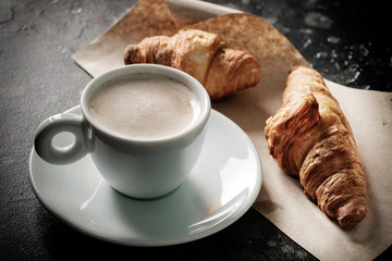 Morning espresso in a cup on a table with croissants behind.