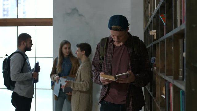 Handsome male college student selecting book from library shelf with group of diverse students talking on background. Clever good looking hipster student looking for a book in university library.