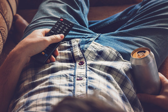 After Work A Guy Lying On Sofa, Drinking A Cold Beer And Watching Sport Tv Channel. Hand Holding A Aluminum Can And Remote Control. Man's Resting Time At Home Concept. 