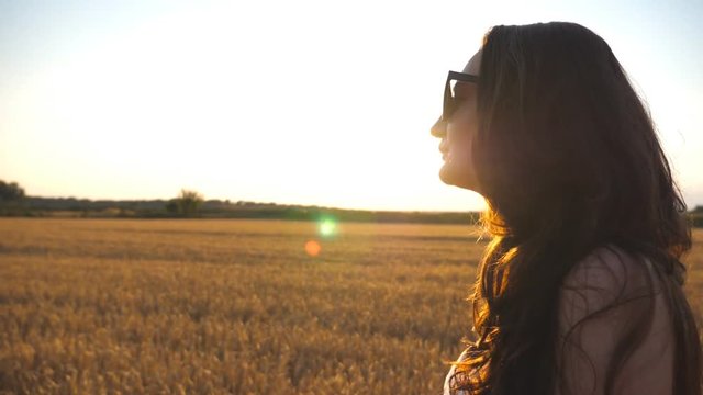 Beautiful girl is walking along wheat field with sun flare at background. Profile of young woman going at the meadow at sunset. Summer leisure at nature concept. Side view Close up Slow motion