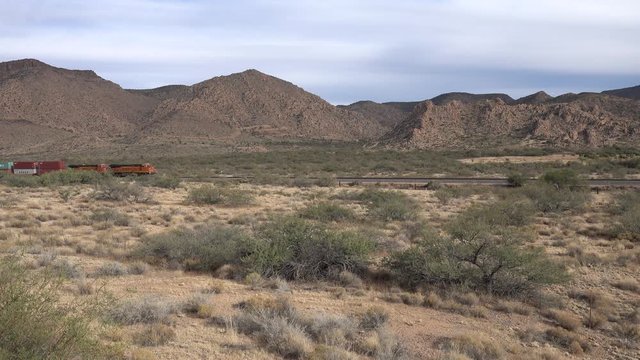A freight train passes from right to left in the desert on a dry hill background. A man walks on a bridge of the locomotive and enters the cabin