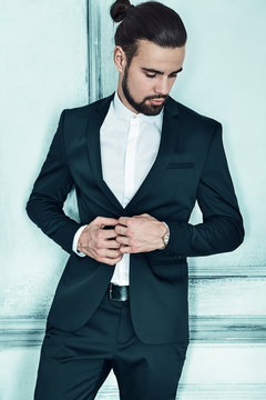 portrait of handsome fashion stylish hipster lumbersexual businessman model dressed in elegant black suit in posing near light blue wall in studio. Metrosexual