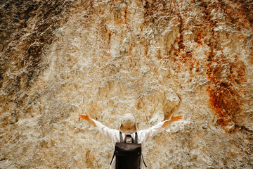 girl with a white shirt and a leather backpack is standing on a stone wall with her hands up. Delight.