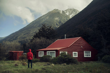 Imagen de un hombre con capucha roja  vigilando delante de una casa roja en medio de la montaña con cielo nuboso. Paisaje de montaña. © josemanuelerre