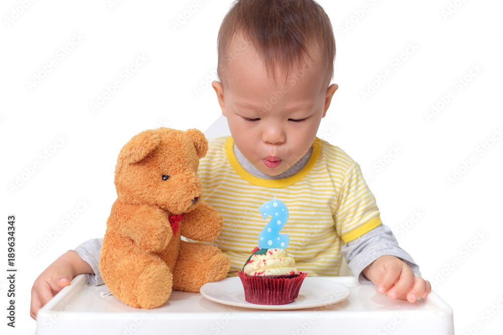 Wall mural Cute little Asian 2  years old toddler baby boy child sitting in high chair celebrating birthday and blowing candle on red velvet cupcake with teddy bear, isolated on white with clipping path