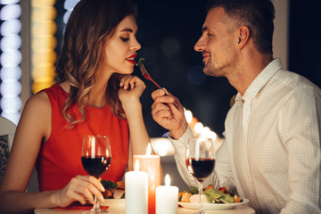Smiling careful man feed his pretty girlfriend while have romantic dinner at home