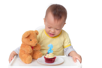 Cute little Asian 2  years old toddler baby boy child sitting in high chair celebrating birthday and blowing candle on red velvet cupcake with teddy bear, isolated on white with clipping path