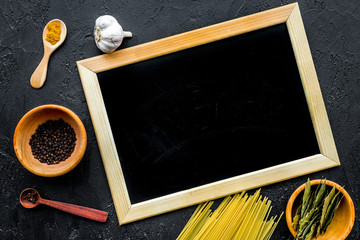 Cook a meal. Mock up for menu or recipe. Chalk board near ingredients. Raw pasta, oil, garlic, spices on black background top view