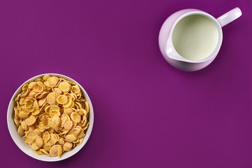 Obraz na płótnie Canvas Bowl with corn flakes, jug of milk on purple background, top view