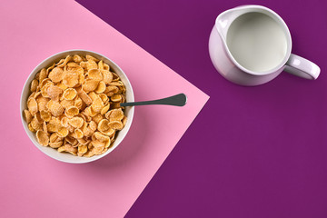 Bowl with corn flakes, jug of milk and spoon on purple and pink background, top view