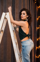 Beautiful curly brunette standing on stairs and posing against lights. Sexy and gorgeous girl wearing in top and jeans with big earrings. Dark loft interior of bedroom.