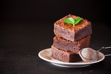 Chocolate brownie square pieces in stack on white plate with walnuts, decorated with mint leaves and cocoa on black background. Delicious dessert. Dark mood. Close up photography. Selective focus