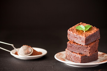 Chocolate brownie square pieces in stack on white plate with walnuts, decorated with mint leaves and cocoa on black background. Delicious dessert. Dark mood. Close up photography. Selective focus