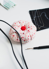 Colorful pin cushion with red button in the middle and stitch thread unpicker or sewing seam ripper with fabric scraps and cables of sewing machine on white crating table