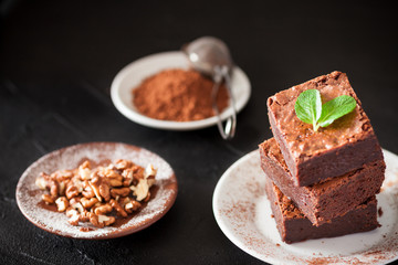 Chocolate brownie square pieces in stack on white plate with walnuts, decorated with mint leaves and cocoa on black background. Delicious dessert. Dark mood. Close up photography. Selective focus